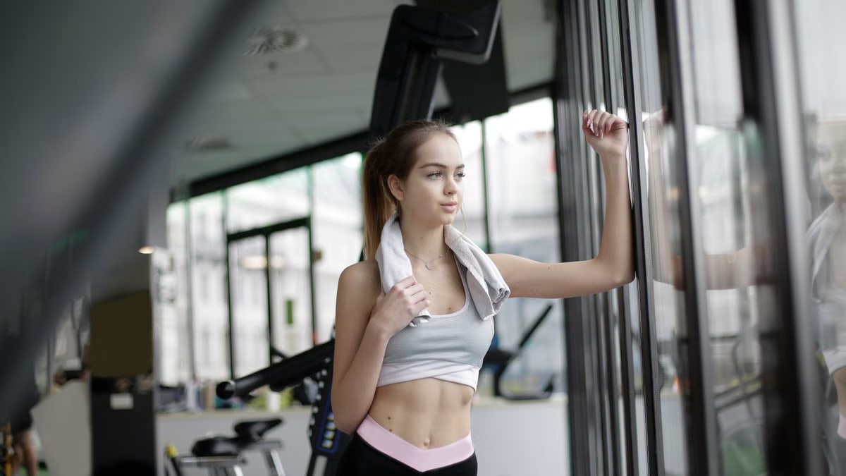 A fit woman stands by a gym window, reflecting after a workout session indoors.